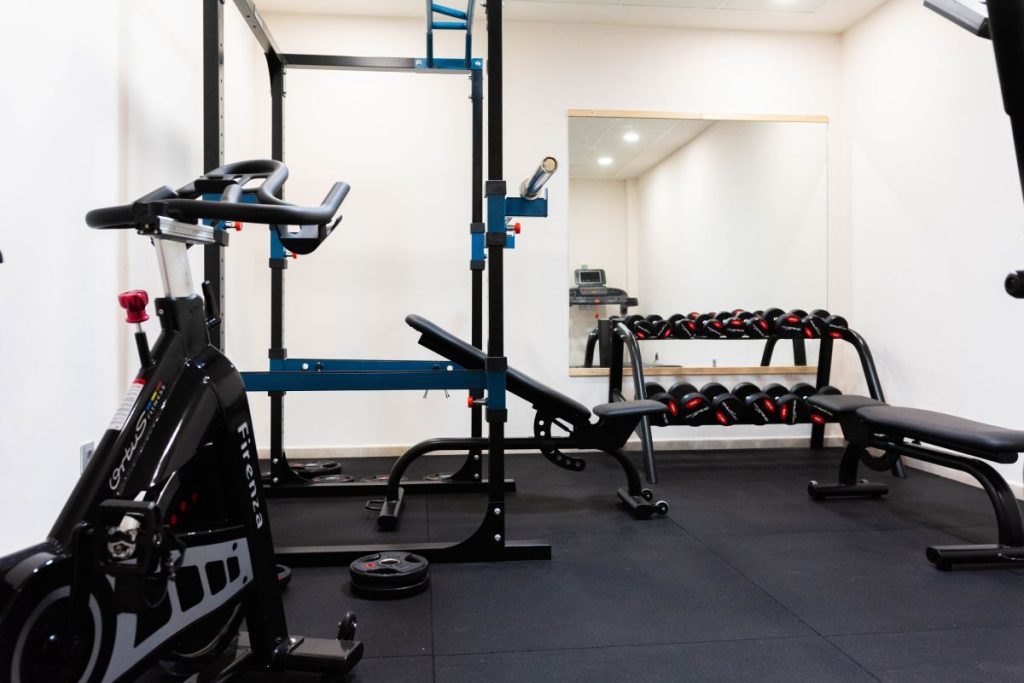 Gimnasio moderno con m&aacute;quina de ejercicio, banco de pesas, pesas y espejo en una sala iluminada.
