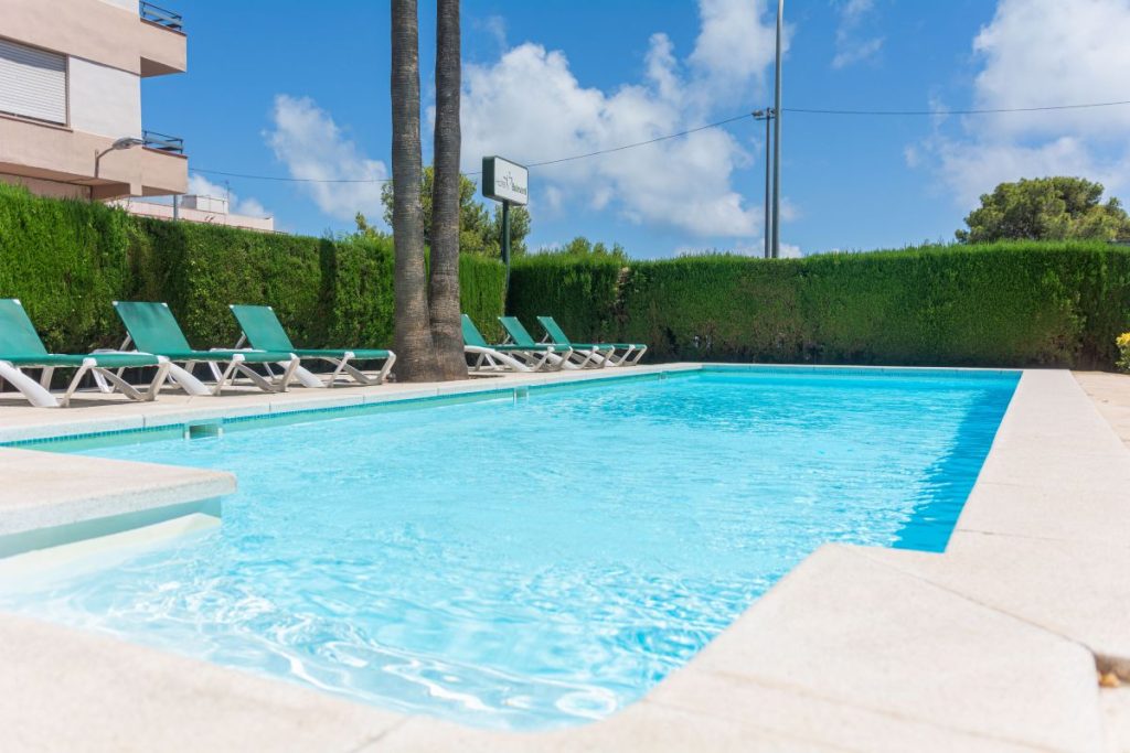Piscina al aire libre con tumbonas verdes, rodeada de setos y palmeras, bajo un cielo azul con nubes.