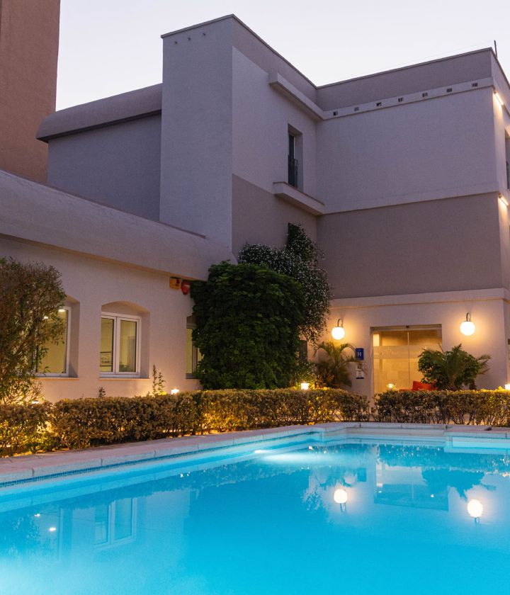 Edificio moderno con piscina iluminada al atardecer, rodeada de arbustos y luces c&aacute;lidas en el jard&iacute;n.