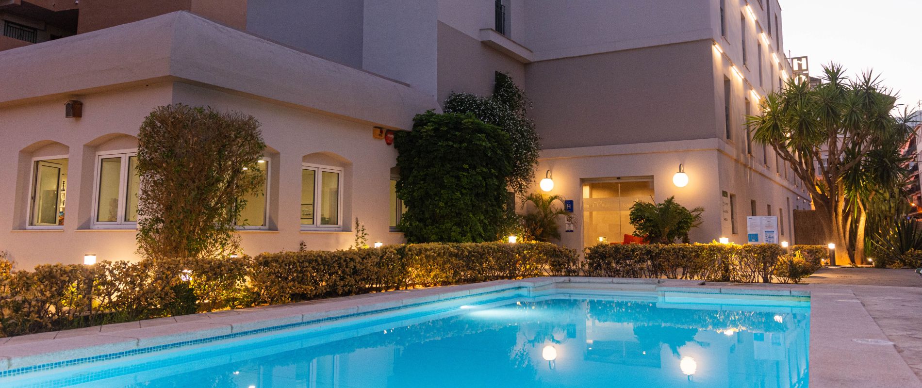 Edificio moderno al atardecer con piscina iluminada, rodeado de plantas y luces exteriores.