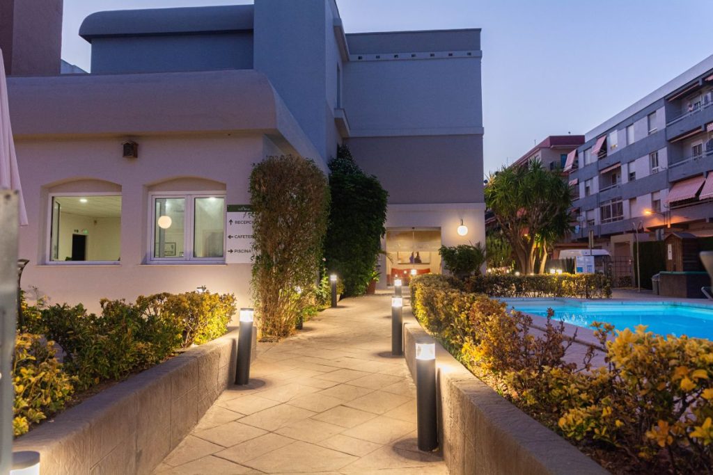 Camino iluminado hacia un edificio moderno, con piscina a la derecha y plantas decorativas a los lados.