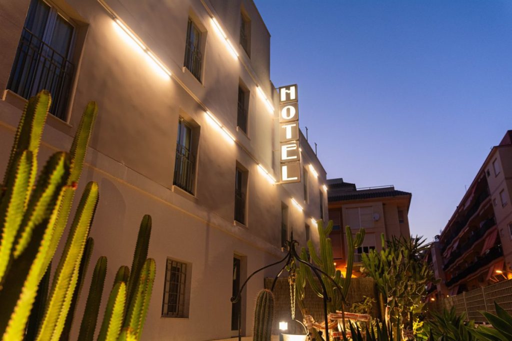 Edificio de hotel iluminado al anochecer, con cactus en primer plano y cielo despejado.